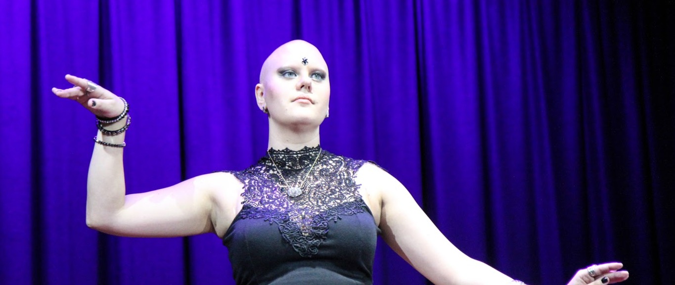 Dominique, a bald white woman is poised mid dance movement. She wears a black lace top and stage makeup. Behind her is a blue curtain.